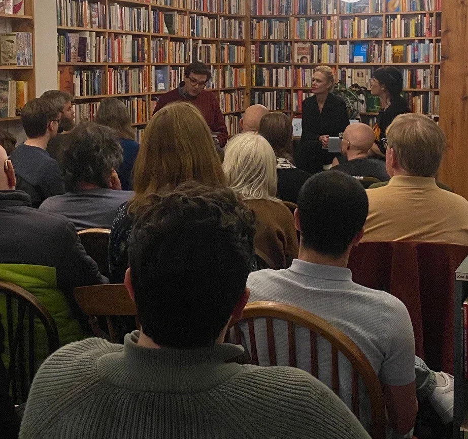 A full audience at an In Other Words evening in Tills Bookshop, seated among the shelves under paper lanterns while three speakers talk at the front